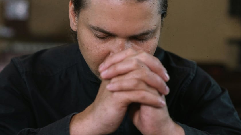 Homem de camisa preta, cabelo preso, de olhos fechados e mãos entrelaçadas diante do rosto.