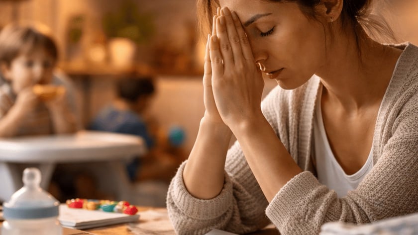 Mulher com as mãos no rosto à mesa, com mamadeira, laptop e crianças ao fundo.