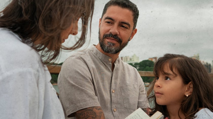 Homem de barba, mulher e menina conversam ao ar livre segurando um livro aberto.