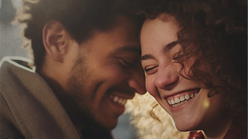 Homem e mulher de cabelos cacheados sorrindo com as testas encostadas, ao ar livre.