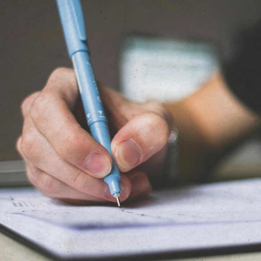 Mão segurando uma caneta azul-clara e escrevendo em um caderno aberto sobre a mesa.