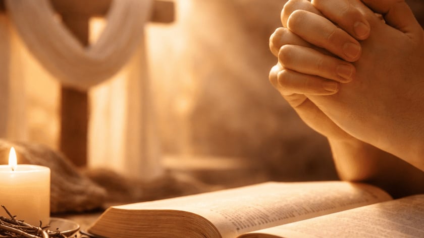 Mãos postas sobre uma Bíblia aberta, perto de uma vela acesa, coroa de espinhos e cruz.