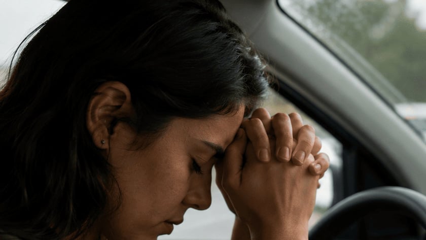 Mulher ao volante de um carro com a testa apoiada nas mãos entrelaçadas, em trânsito parado.