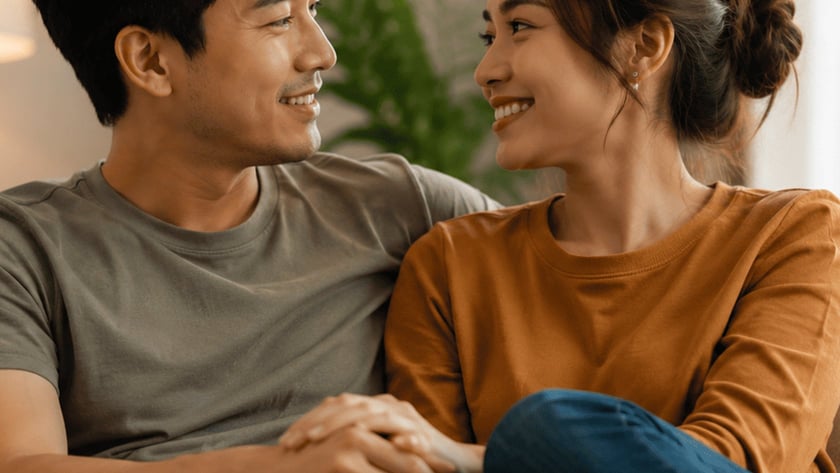 Casal sentado no sofá, de mãos dadas, olhando um para o outro e sorrindo.