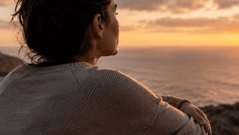 Mulher de coque e suéter bege, vista de costas, observa o mar do alto de um penhasco.