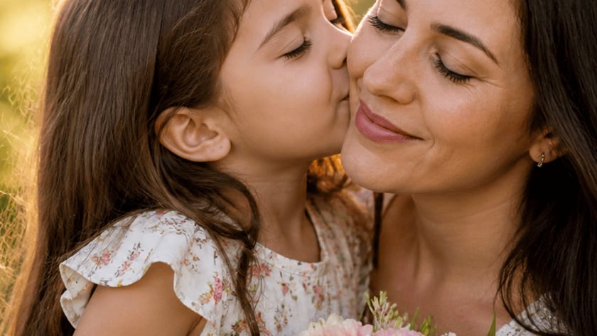 Menina de cabelos castanhos beija a bochecha de uma mulher que segura um buquê de flores rosa.