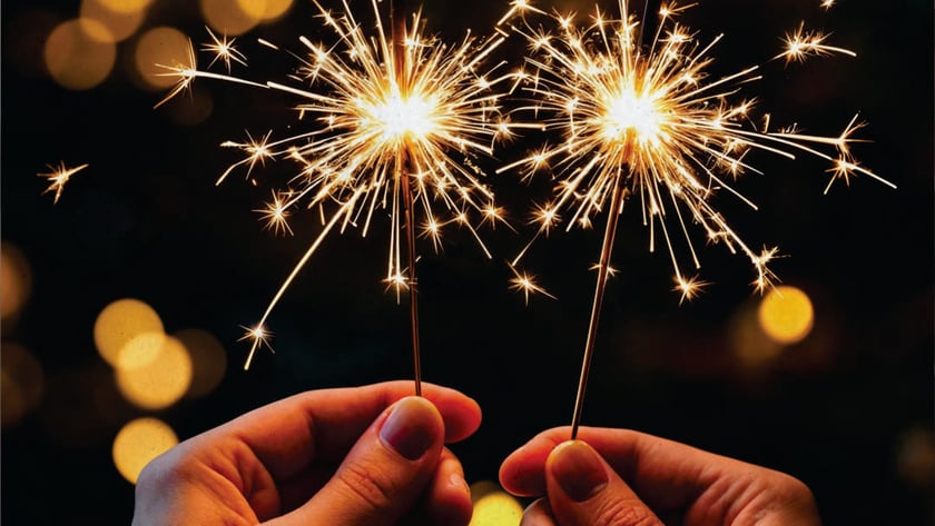 Duas mãos seguram estrelinhas acesas, soltando faíscas douradas à noite.