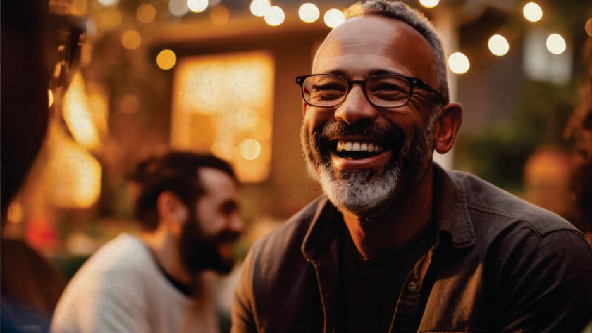 Homem de óculos e barba grisalha rindo em um jardim com luzes ao fundo.