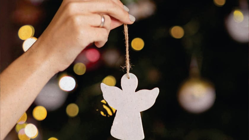 Mão segura enfeite de anjo branco pendurado por barbante diante de árvore de Natal.