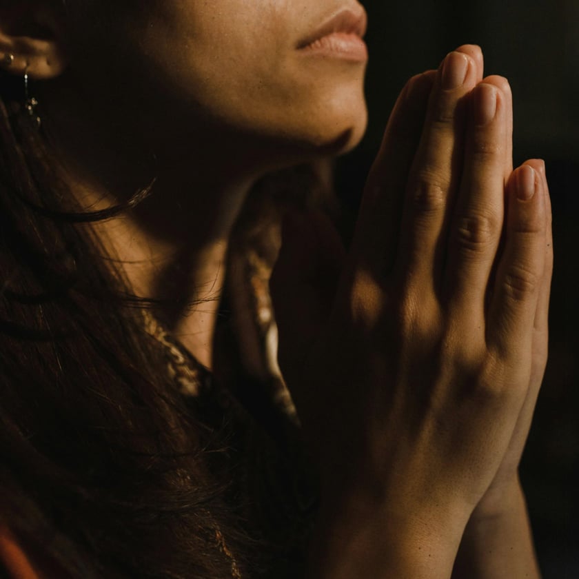 Mulher de cabelos escuros com as mãos unidas diante do queixo, em um ambiente escuro.