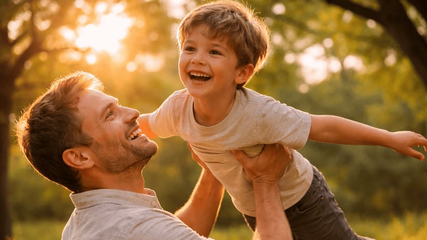 Homem sorridente levanta um menino com os braços abertos, ao ar livre.