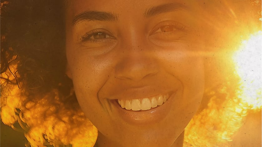 Mulher de cabelo cacheado sorri para a câmera, com o sol atrás dela.