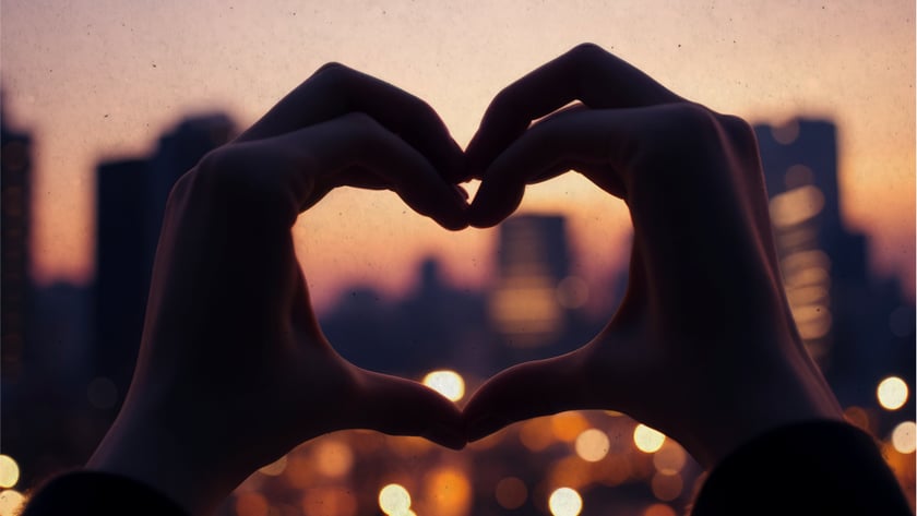 Duas mãos formam um coração diante de uma cidade ao entardecer, com luzes desfocadas.