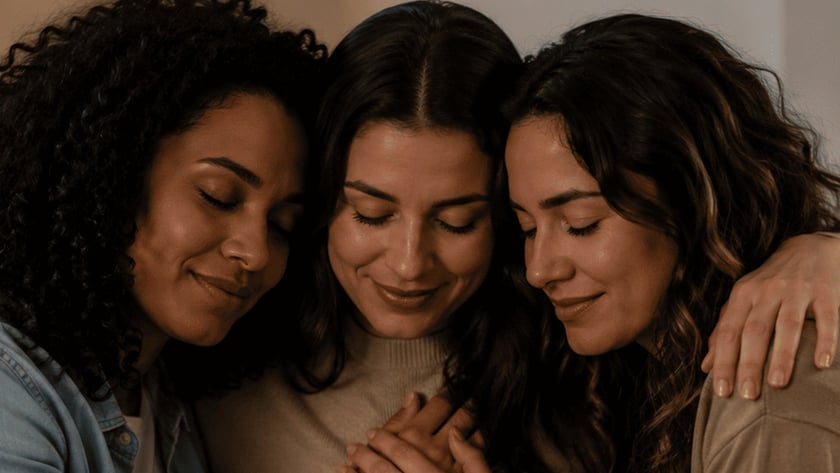 Três mulheres abraçadas com as cabeças encostadas e as mãos unidas sobre o peito da do meio.