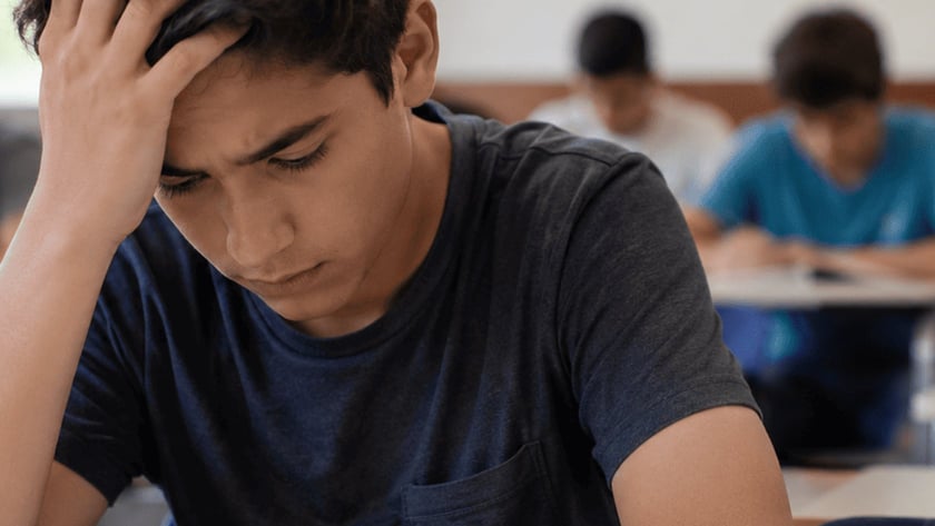 Estudante de cabelo escuro, com a mão na testa, olha para um caderno em sala de aula.