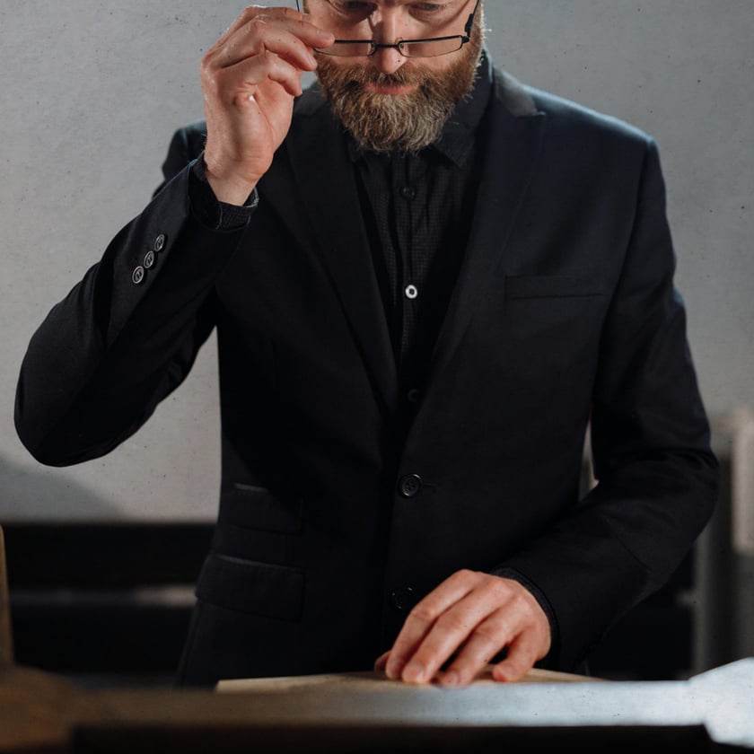 Homem barbudo de terno preto ergue os óculos e olha papéis sobre uma mesa.