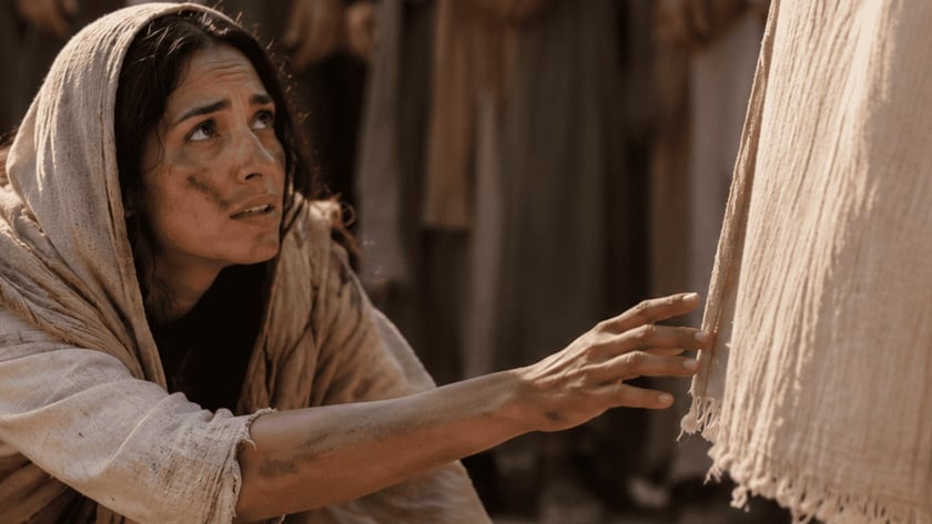Mulher ajoelhada na terra estende a mão para tocar a barra de uma túnica clara.