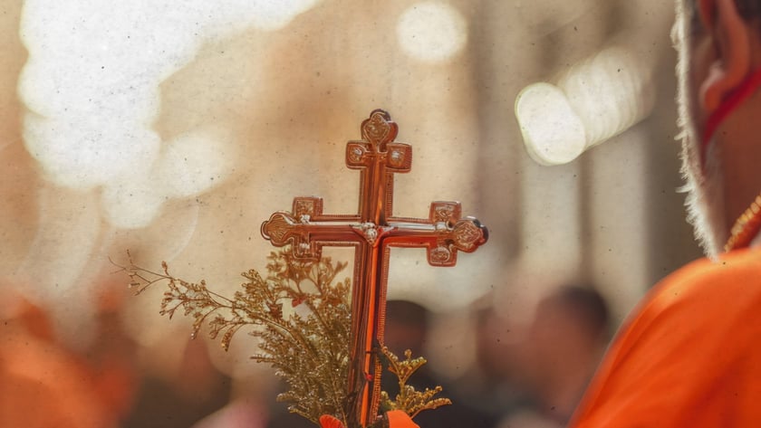Religioso de vestes laranja segura uma cruz dourada com ramos e fita, diante de uma multidão.