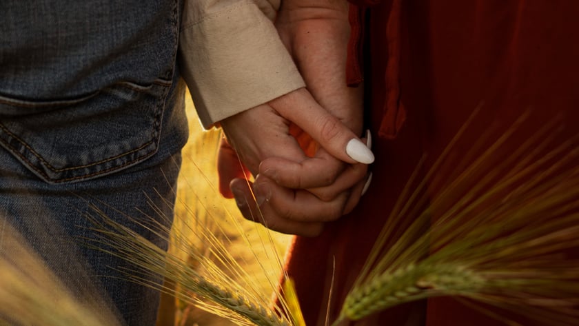 Duas pessoas de mãos dadas em um campo de trigo, uma de jeans e outra de vestido vermelho.