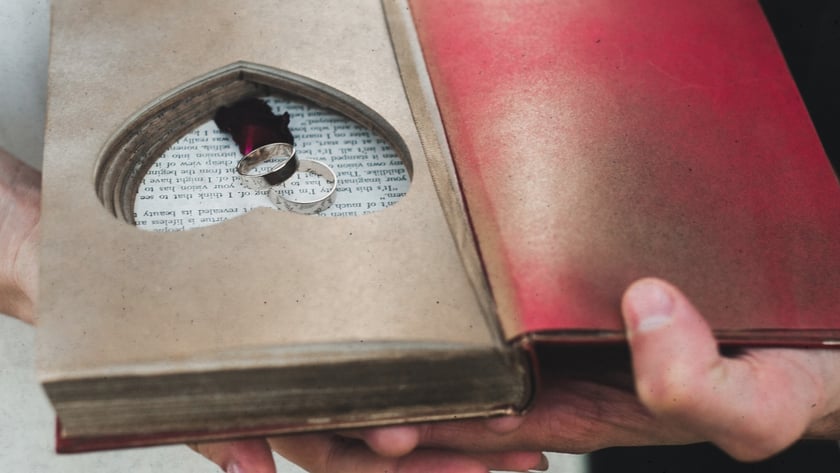 Duas pessoas seguram um livro aberto com recorte em coração contendo duas alianças de prata.