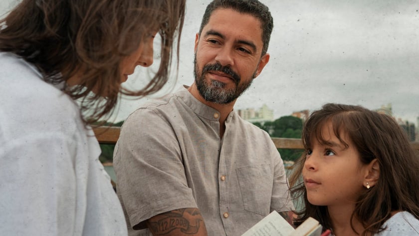 Homem de barba, mulher e menina conversam ao ar livre segurando um livro aberto.