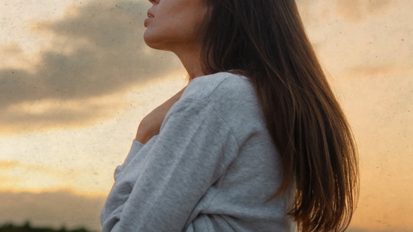 Mulher de perfil, olhos fechados, mão no peito, com moletom cinza ao entardecer.
