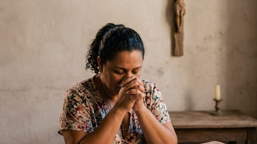 Mulher de olhos fechados e mãos juntas reza diante de uma Bíblia aberta, com crucifixo na parede.