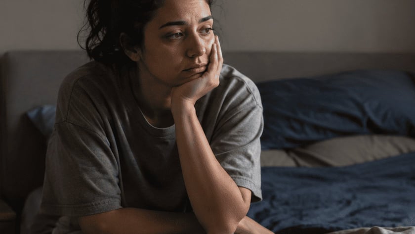 Mulher de camiseta cinza sentada na beira de uma cama desarrumada, com a mão no rosto.