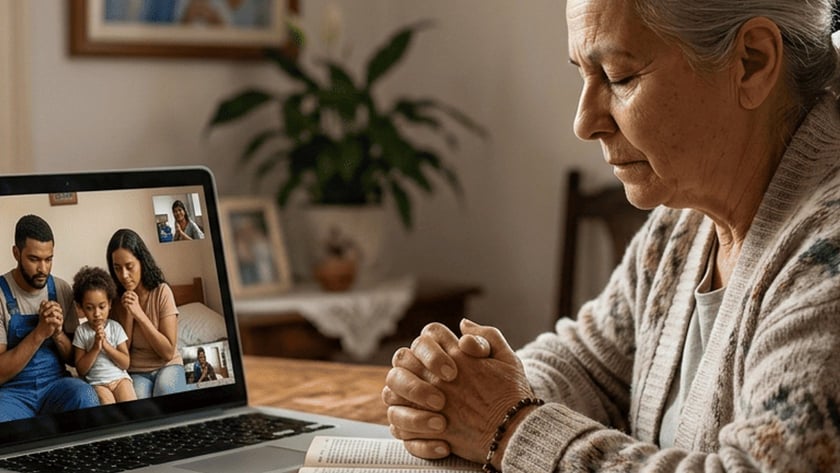 Idosa de mãos postas sobre uma Bíblia reza diante de um notebook com videochamada de família.