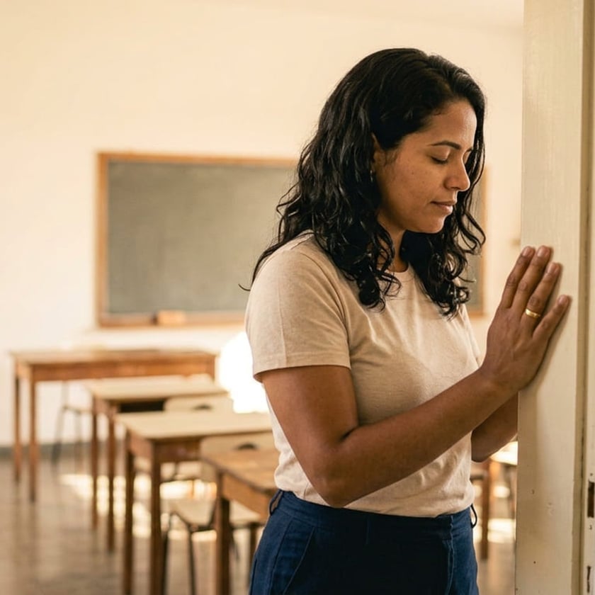 Mulher de cabelo escuro e olhos fechados apoia a mão na parede de uma sala de aula vazia.