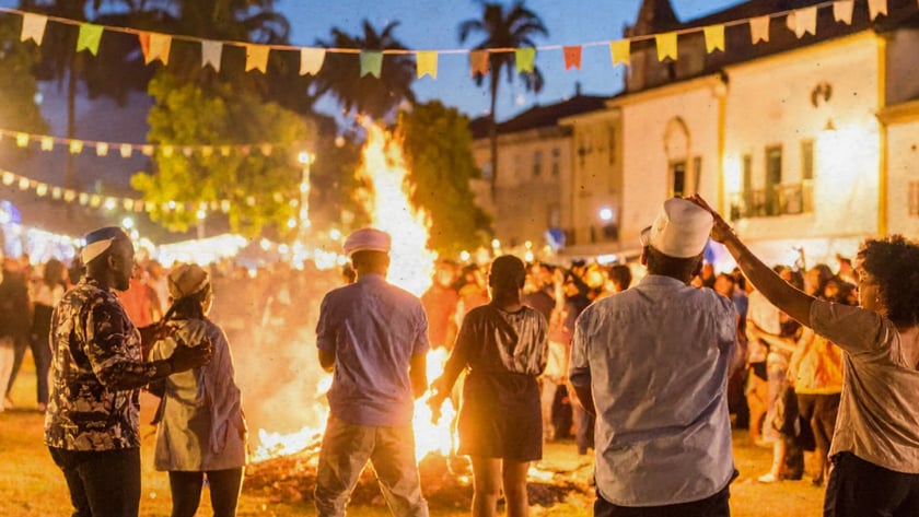 Pessoas de costas dançam ao redor de uma fogueira numa praça com bandeirinhas coloridas.