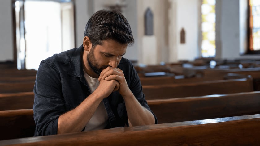 Homem de barba, com as mãos unidas junto ao rosto, sentado em um banco de igreja.