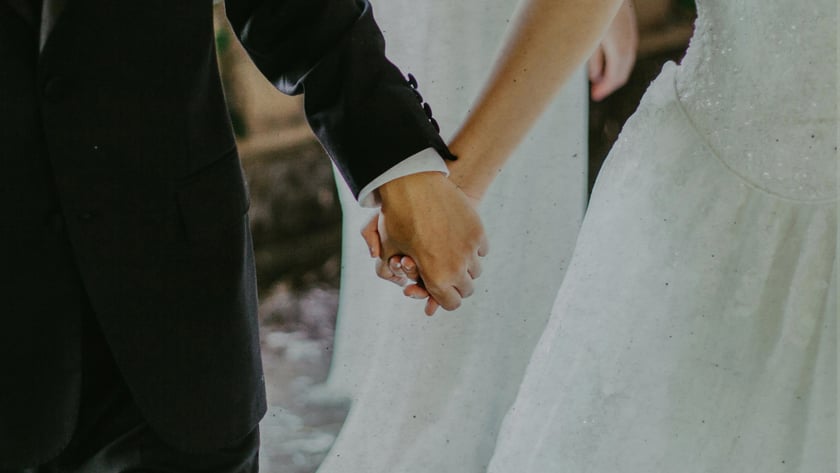 Noiva de vestido branco e noivo de terno escuro andam de mãos dadas.