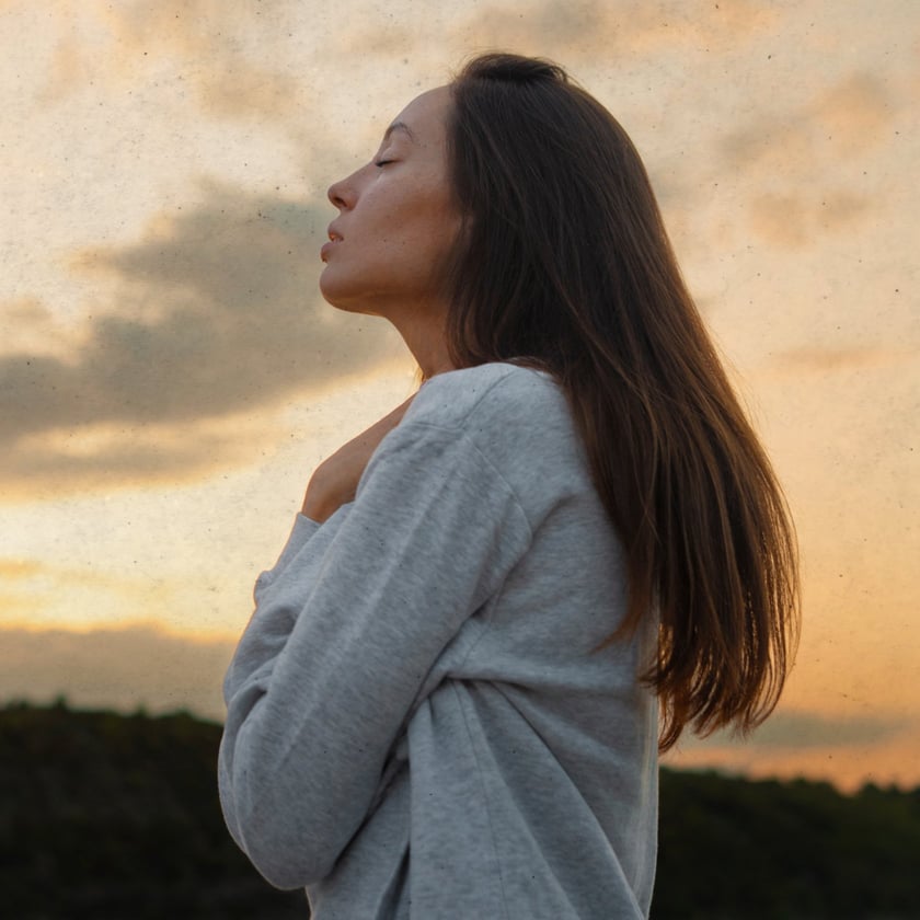 Mulher de perfil, olhos fechados, mão no peito, com moletom cinza ao entardecer.