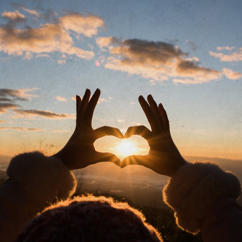 Mãos de uma pessoa formam um coração em torno do sol nascente sobre montanhas.