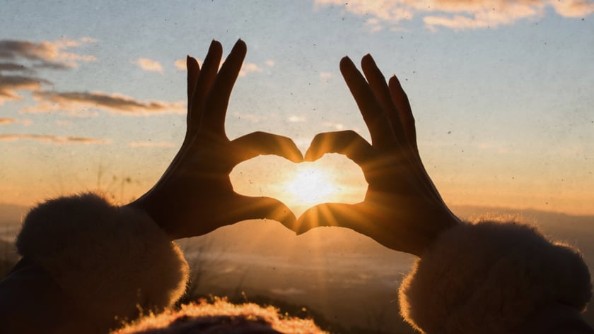 Mãos de uma pessoa formam um coração em torno do sol nascente sobre montanhas.