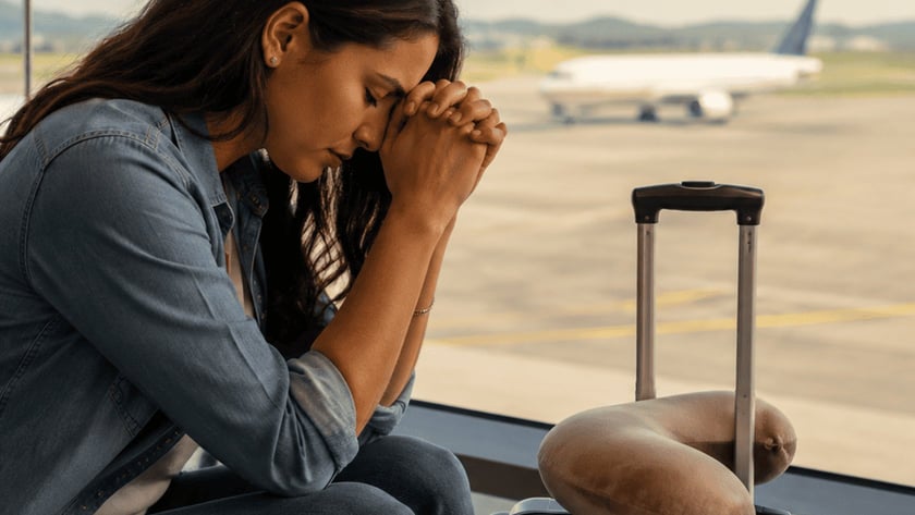 Mulher de cabelo escuro, com as mãos juntas na testa, sentada em aeroporto ao lado de mala.