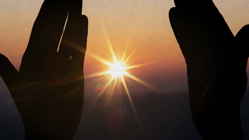 Duas mãos erguidas em silhueta enquadram o sol brilhando no horizonte ao entardecer.