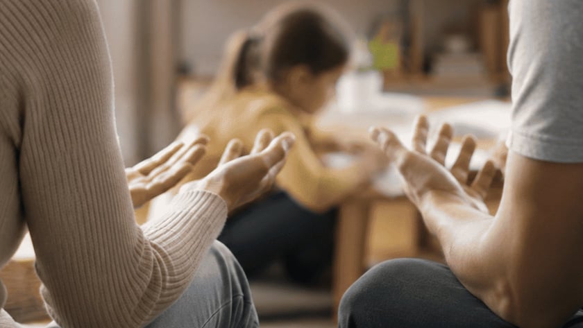 Duas pessoas sentadas num sofá com as palmas das mãos abertas; ao fundo, uma criança brinca.