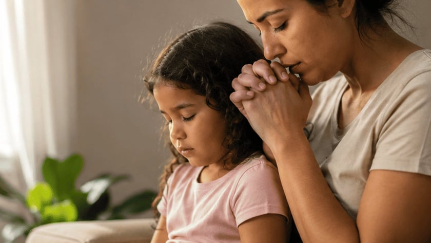 Mulher e menina de olhos fechados e mãos juntas, rezando no sofá com uma Bíblia aberta.
