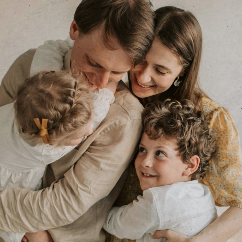 Pai, mãe, menino de cabelo cacheado e menina pequena se abraçando diante de uma parede clara.