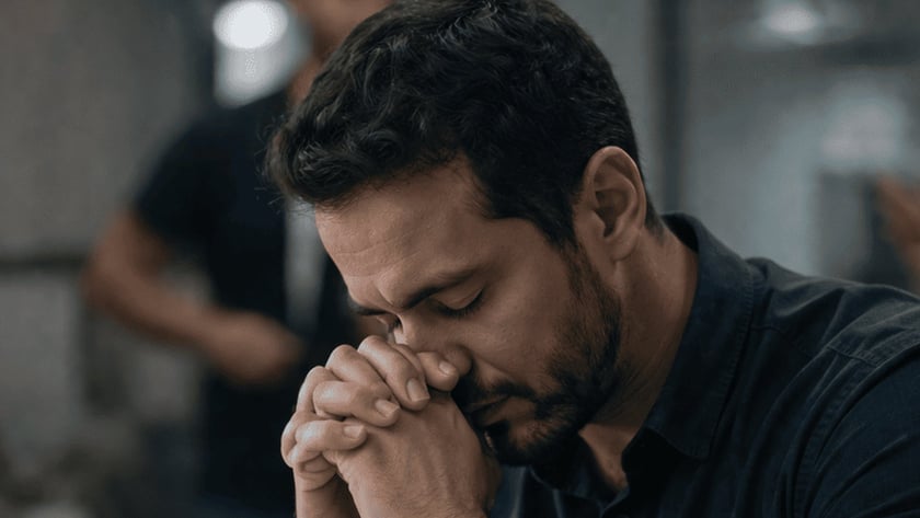Homem de mãos postas e olhos fechados diante de um laptop e de um livro aberto.