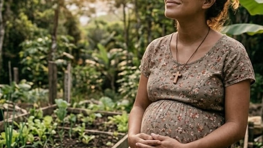 Mulher grávida sentada em horta cercada de mata, com Bíblia no colo e xícara ao lado.