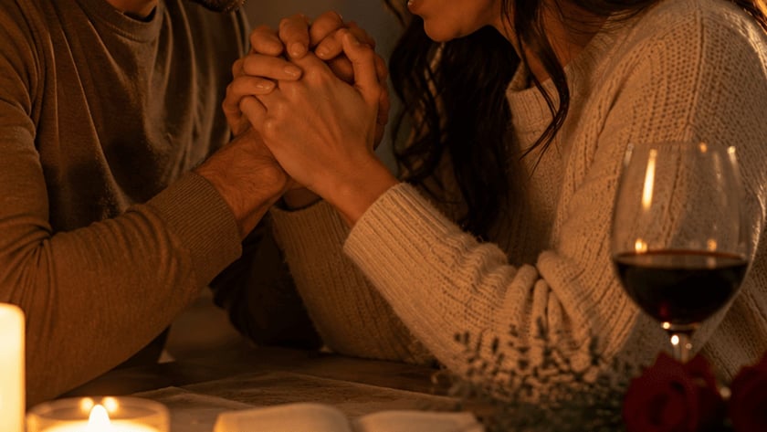 Casal sentado à mesa, de olhos fechados, com as testas encostadas e as mãos unidas.