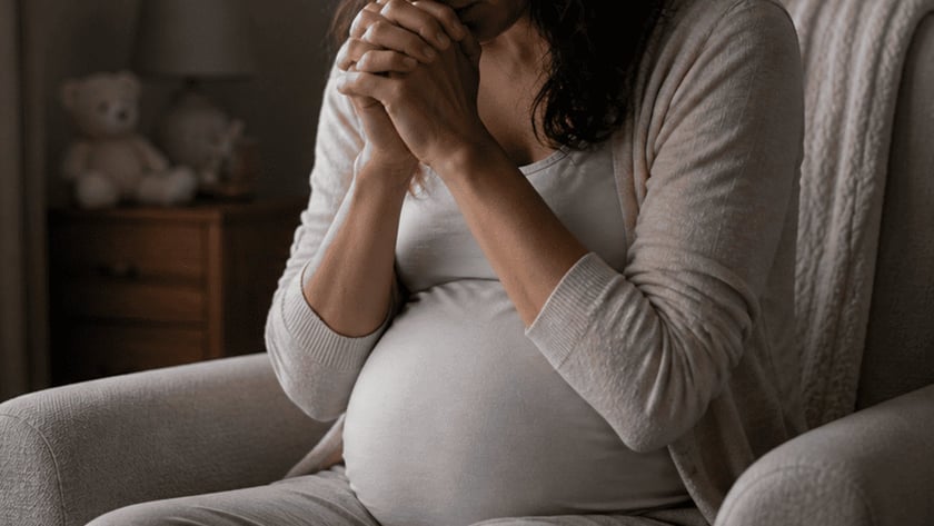 Mulher grávida sentada em poltrona com as mãos unidas junto ao rosto, de olhos fechados.