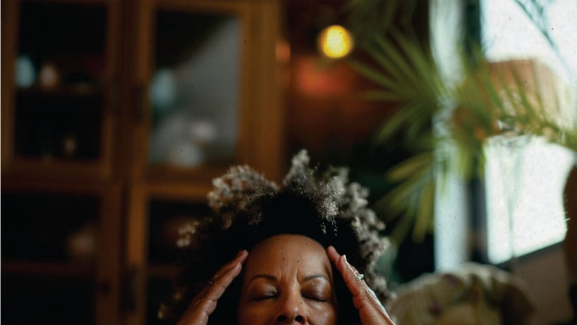 Mulher de olhos fechados pressiona as têmporas com as mãos, sentada num sofá na sala.