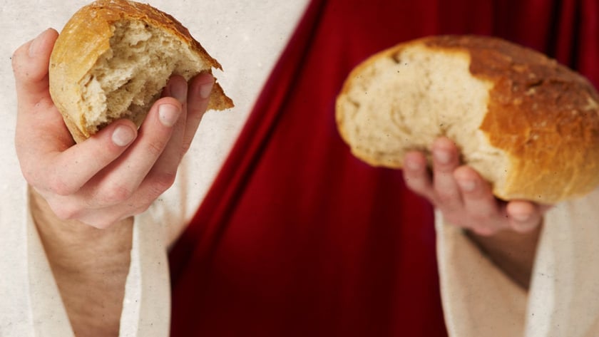 Pessoa de túnica clara e manto vermelho segura dois pedaços de pão partido.