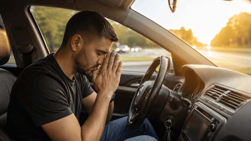 Homem de camiseta escura sentado ao volante, com as mãos unidas diante do rosto e olhos fechados.