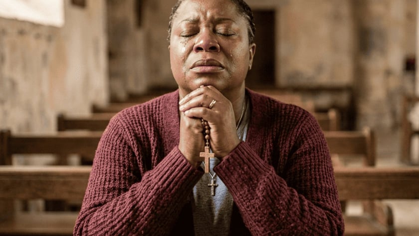 Mulher de cardigã vinho reza com um terço nas mãos em um banco de igreja.