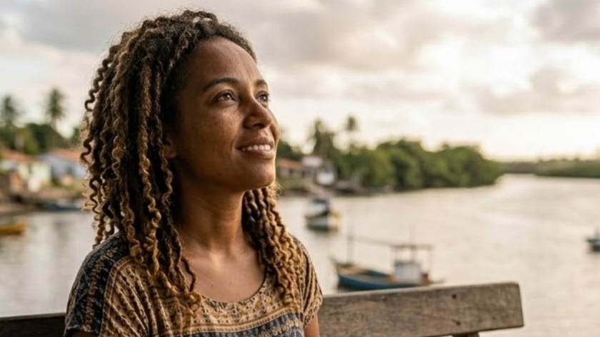 Mulher de cabelo cacheado sentada num banco de madeira com um livro aberto, diante de um rio.